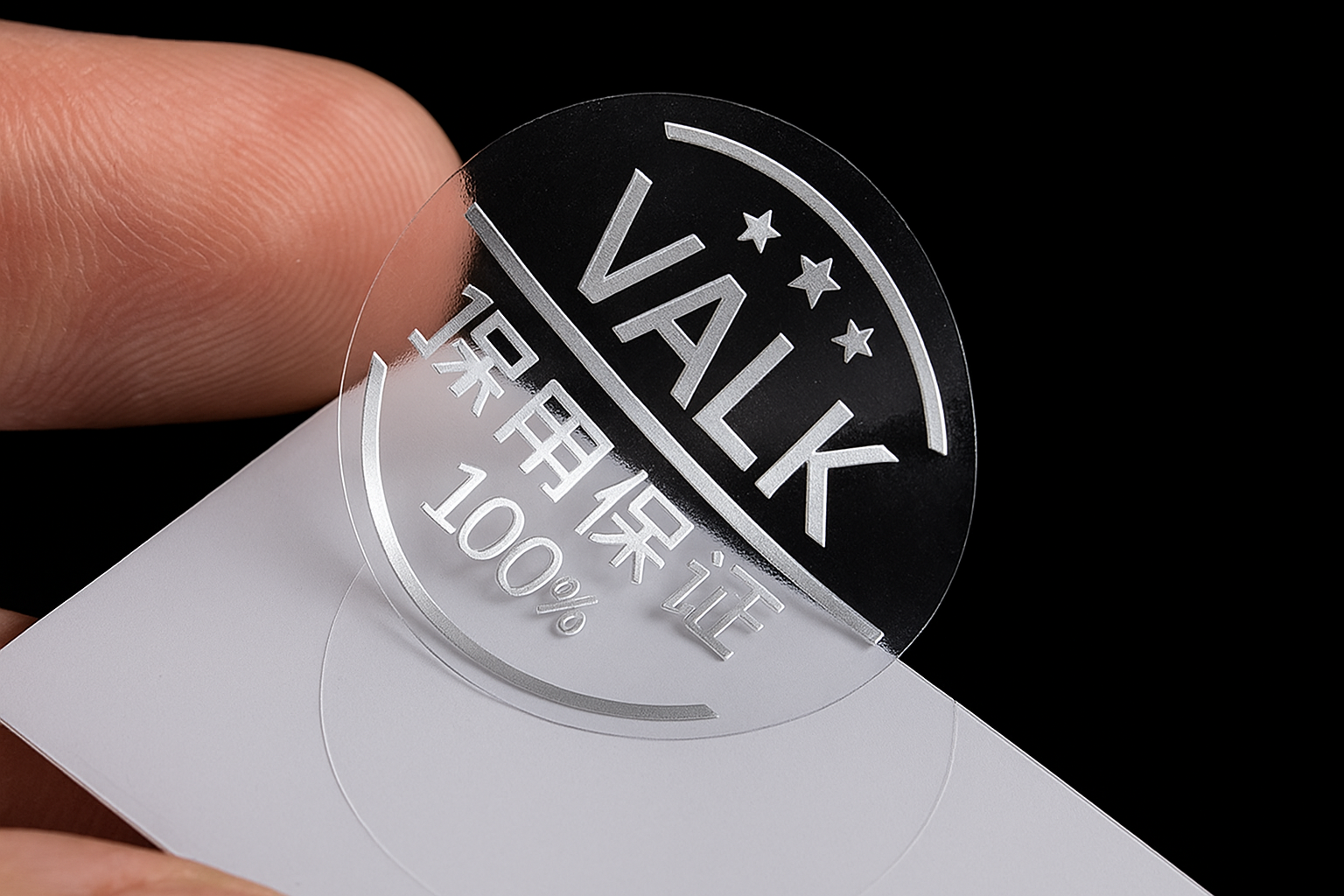 Un adesivo trasparente argentato circolare con un logo, tenuto da un dito contro uno sfondo nero, che mostra la qualità trasparente e riflettente del materiale